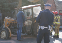 Umundurowany policjant wykonuje oględziny uszkodzonego w wyniku zdarzenia drogowego ciągnika rolniczego. Fotografia poglądowa.