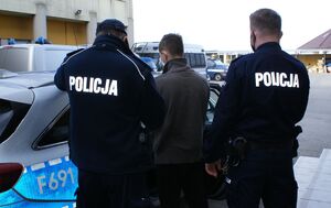 Umundurowani policjanci eskortują zatrzymanego do radiowozu.