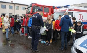 Policjanci, strażacy, ratownicy medyczni oraz przedstawiciele samorządu podczas spotkania profilaktycznego z młodzieżą. W tle radiowozy poszczególnych służb.