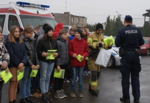 Policjanci, strażacy, ratownicy medyczni oraz przedstawiciele samorządu podczas spotkania profilaktycznego z młodzieżą. W tle radiowozy poszczególnych służb.