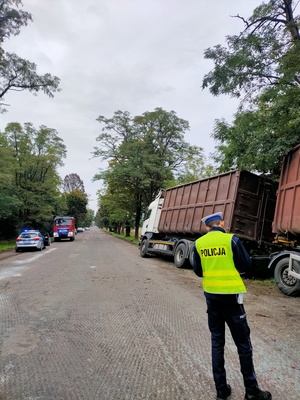 Na zdjęciu widoczny policjant ruchu drogowego wykonujący swoje czynności na tle zespołu pojazdów ( ciężarowa scania wraz z przyczepą), z lewej strony  widać radiowóz policyjny oraz samochód strażacki.