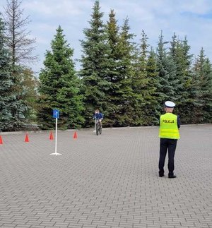 widzimy policjanta egzaminującego ucznia jadącego rowerem i pokonującego tor przeszkód.