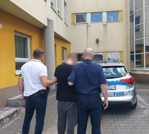 policjanci prowadza zatrzymanego, w tle widzimy budynek komendy i oznakowany radiowóz.