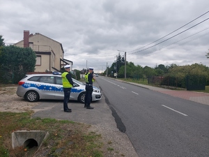 widzimy policjantów stojących przy radiowozie, jeden z nich mierzy prędkość jadącego samochodu.