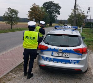 widzimy policjantów mierzących prędkość pojazdu, obok stoi radiowóz.