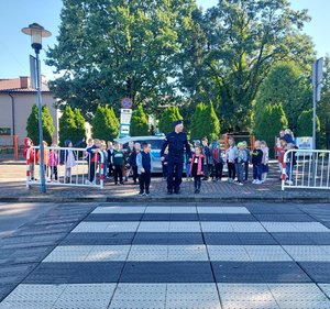 widzimy policjanta prowadzącego po przejściu dla pieszych dwójkę dzieci. W tle widać radiowóz i pozostałe dzieci.