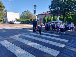 widzimy policjanta prowadzącego po przejściu dla pieszych dzieci. W tle widać radiowóz i pozostałe dzieci.