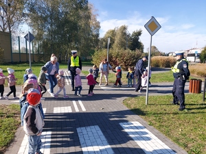 widzimy policjanta, który kieruje ruchem drogowym na miasteczku ruchu drogowego, a dzieci prowadzone są przez innego policjanta.