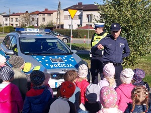 widzimy policjantów z dziećmi stojących przy radiowozie na którym znajdują się elementy odblaskowe.