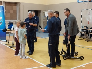 widzimy policjanta wręczającego medale uczniom w tle widać inne osoby.