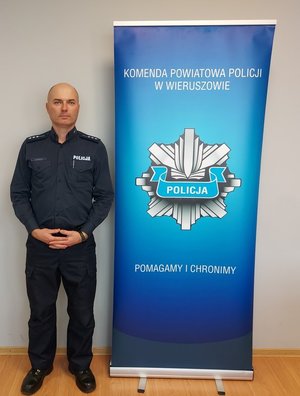 widzimy policjanta stojącego w pomieszczeniu obok baneru z napisem Policja Wieruszów. Pomagamy i chronimy.