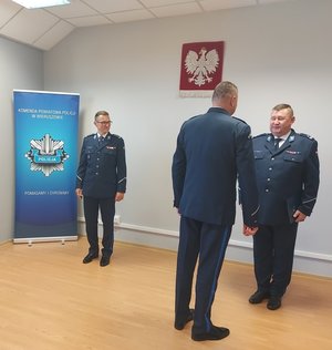 Widzimy w pomieszczeniu Zastępce Komendanta Wojewódzkiego Policji w Łodzi inspektora Andrzeja Patrzałek gratulujący obejmującemu stanowisko I Zastępcy Komendanta Powiatowego Policji w Wieruszowie podinspektorowi Pawłowi Mosce. Obok stoi Komendant Powiatowy Policji w Wieruszowie inspektor Grzegorz Dzierzkowski. W tle widać rolap komendy i policjanta.