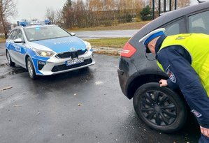 widzimy policjanta sprawdzajacego opony w samochodzie w tle widzimy radiowóz policyjny.