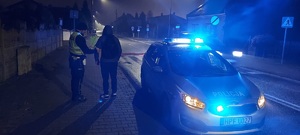 widzimy policjanta wręczającego element odblaskowy mężczyźnie na tle radiowozu z włączonymi światłami błyskowymi.