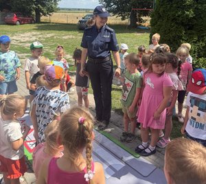 widzimy dzieci podczas spotkania z policjantami, które prowadzone jest na placu przedszkola. Policjantka omawia zasady ruchu drogowego na makiecie edukacyjnej.
