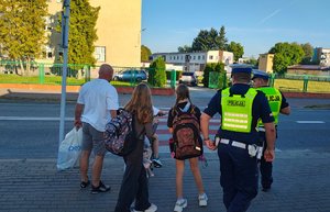 widzimy policjantów prowadzących dzieci wraz z opiekunem do przejścia dla pieszych.