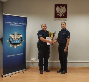 widzimy Komendanta, który gratuluje policjantowi trzymający puchar oraz dyplom.