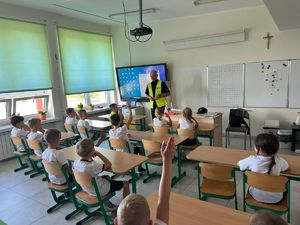 widzimy policjanta ubranego w kamizelkę odblaskową. Zajęcia są w klasie a uczniowie siedzą w ławkach.
