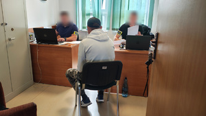 widzimy zatrzymanego mężczynę oraz policjantów pracujących w pokoju siedzących przy biurkach.