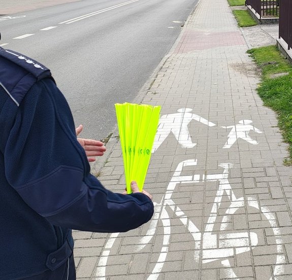 widzimy jak policjant trzyma odblaski w ręku na tle drogi i ścieżki rowerowej.