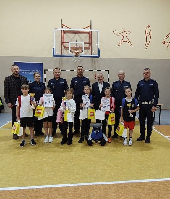 widzimy laureatów konkursu wraz z organizatorami i zaproszonymi gośćmi. Zdjęcie jest na sali gimnastycznej a dzieci trzymają w ręku nagrody w postaci pucharów, dyplomów i nagród rzeczowych.