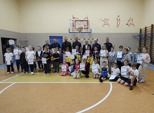 widzimy zdjęcie grupowe wszystkich uczestników konkursu wraz z organizatorami i zaproszonymi gośćmi. Zdjęcie jest na sali gimnastycznej a dzieci trzymają w ręku dyplomy oraz nagrody w postaci pucharów, dyplomów i nagród rzeczowych.