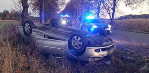 widzimy uszkodzony pojazd leżący na dachu oraz policjanta, strażaka oraz w tle karetkę pogotowia.