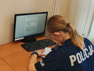 widzimy policjantkę siedzącą przy biurku, przed komputerem piszącą w notatniku służbowym.