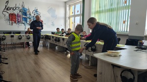 widzimy policjantów prowadzących spotkanie, oraz policjantkę, która pomaga uczniowi założyć kamizelkę odblaskową.