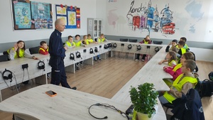 widzimy policjantów prowadzących spotkanie oraz uczniów siedzących w ławkach ubranych w kamizelki odblaskowe.