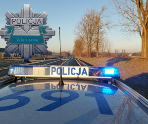 widzimy belkę policyjnego radiowozu na tle drogi oraz widzimy wklejona z lewej strony odznakę policyjna z napisem Policja Wieruszów.