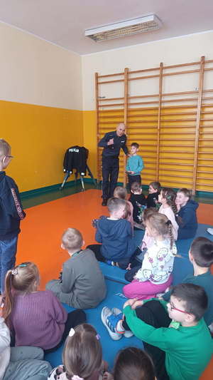 widzimy policjanta prowadzącego spotkanie z dziećmi siedzących w sali.