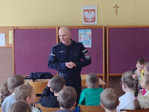 widzimy policjanta prowadzącego spotkanie z dziećmi siedzących w sali.