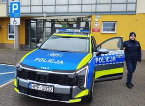 widzimy policjantkę stojąca przy radiowozie na tle budynku komendy.