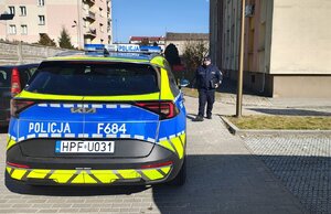widzimy policjanta idącego między blokami w tle widać policyjny radiowóz.