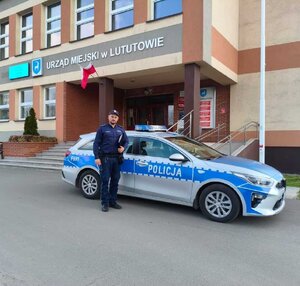 widzimy policjanta stojącego przy radiowozie przed budynkiem gminy.
