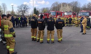 widzimy odprawę służb na parkingu widzimy policjantów strażaków oraz pojazdy służbowe.