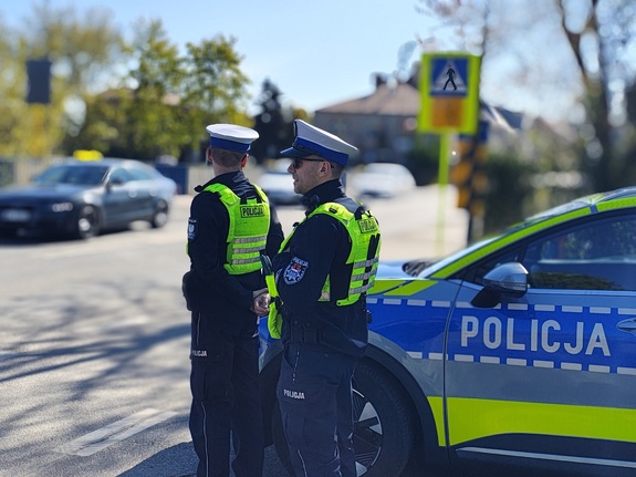 widzimy policjantów stojących przy radiowozie oraz przejeżdżające pojazdy.