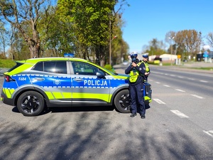 widzimy policjanta mierzącego prędkość, a drugu funkcjonariusz stoi obok policyjnego pojazdu.