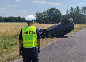 widzimy policjanta na tle uszkodzonego samochodu leżącego na dachu.