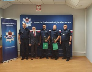 widzimy policjantów w auli komendy na tle baneru niebieskiego koloru z napisem Komenda Powiatowa Policji w Wieruszowie. Mundurowi w rękach trzymają teczki.
