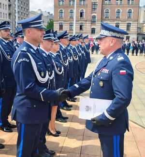 widzimy policjanta gratulującego innemu policjantowi oraz szereg policjantów stojących w rzędzie.