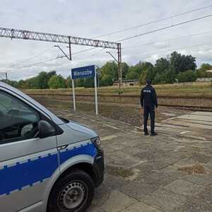 widzimy policjanta stojącego przy przejeździe kolejowym, widzimy tez ustawiony policyjny radiowóz.