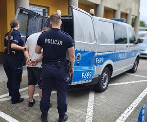 widzimy mężczyznę prowadzonego przez policjantów do radiowozu typu bus.