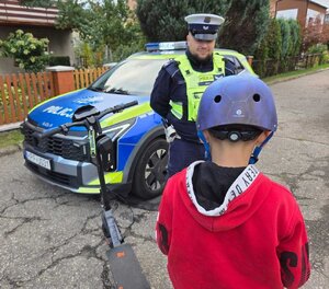 widzimy policjanta i chłopca w kasku obok hulajnoga i policyjny radiowóz.