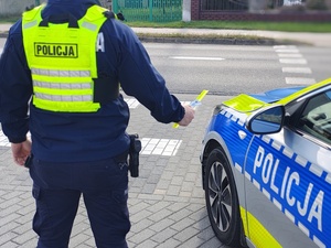 widzimy policjanta stojącego przy radiowozie trzymającego odblaski w ręku.